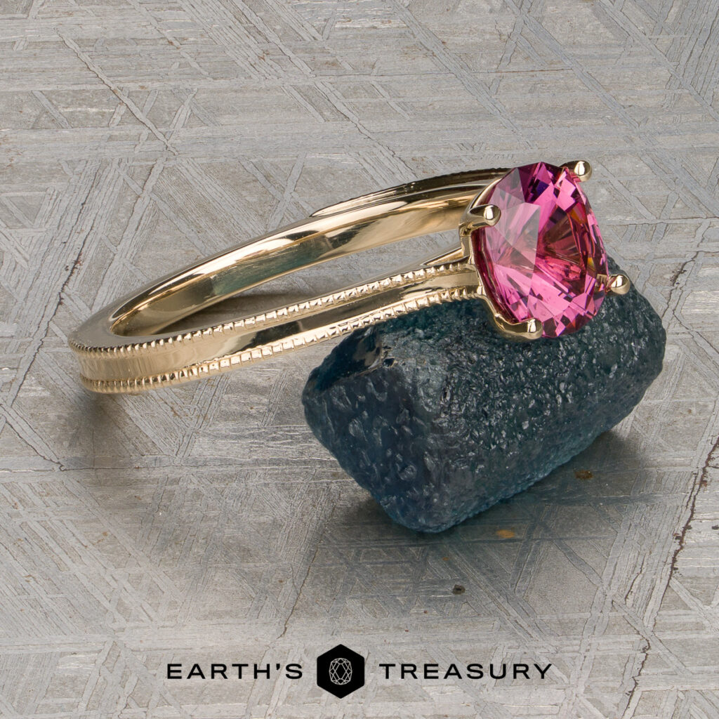 A gold ring with intricate filigree and a pink gemstone sits atop a rugged dark blue stone, displayed on a patterned metallic surface. The image is stacked from 46 images (Method=B, R=7, S=2). "EARTH'S TREASURY" logo and text are visible at the bottom of the image.