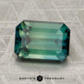 A rectangular, emerald-cut 3.23-carat Madagascar sapphire (heated) in a vivid blue-green hue rests on a textured gray surface, with the text "EARTH'S TREASURY" visible below the gem.