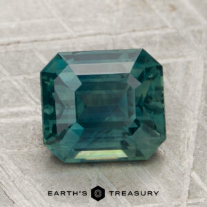 A 3.03-carat, rectangular emerald-cut blue-green sapphire (heated) is displayed on a textured gray surface with the "Earth's Treasury" logo below.