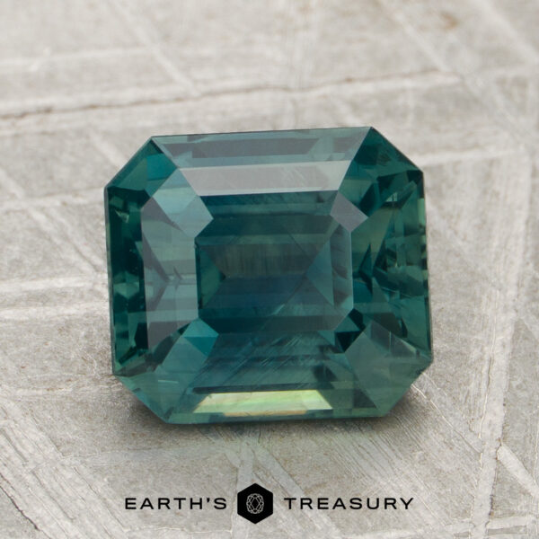 3.03-Carat Sapphire (Heated) A 3.03-carat, rectangular emerald-cut blue-green sapphire (heated) is displayed on a textured gray surface with the "Earth's Treasury" logo below.