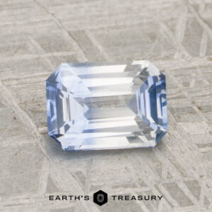 A rectangular, faceted 1.56-carat heated sapphire with a light blue hue is displayed on a textured gray surface, featuring the "Earth's Treasury" logo and text at the bottom.