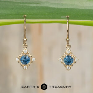 Stacked from 16 images using Method=B (R=7, S=2), these gold drop earrings feature blue center gemstones with white accent stones and are displayed hanging from a green leaf. The "EARTH'S TREASURY" logo and text appear at the bottom of the image.