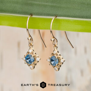 Stacked from 20 images (Method=B, R=7, S=2) gold drop earrings with blue gemstones and small clear stones are displayed hanging from a green leaf. The Earth's Treasury logo and name appear at the bottom of the image.