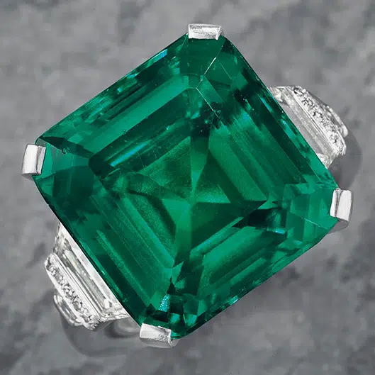 A ring named Screen-Shot-2017-05-16-at-7.46.47-AM-e1494958828147, featuring a square-cut green emerald inspired by the Emerald Guide, is set in a silver band with small diamonds on the sides and displayed on a gray textured background.