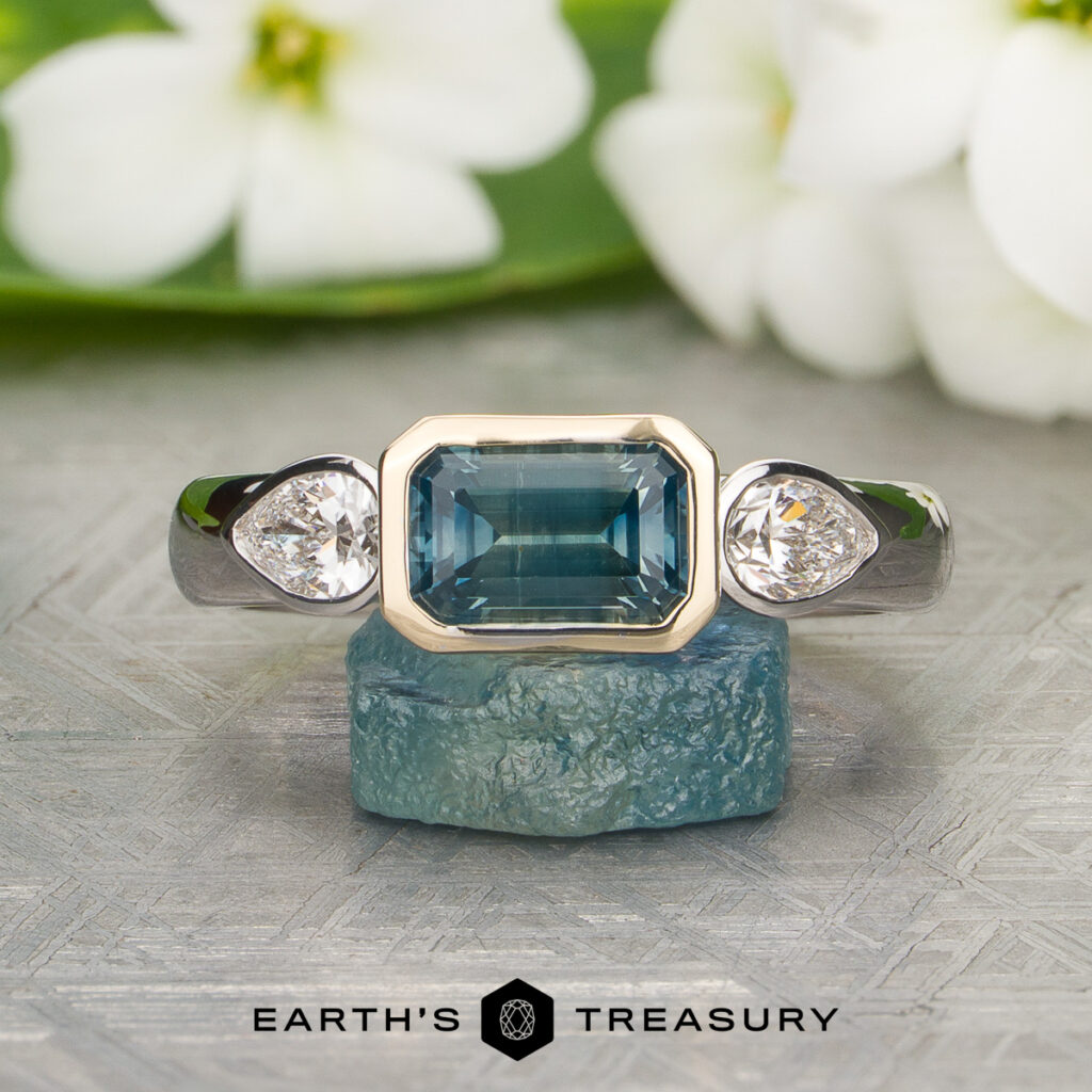 Susan Gabor [custom gallery] rectangular blue gemstone ring with two side diamonds, displayed on a blue stone and white flowers in the background. The logo text "Earth's Treasury" appears at the bottom.
