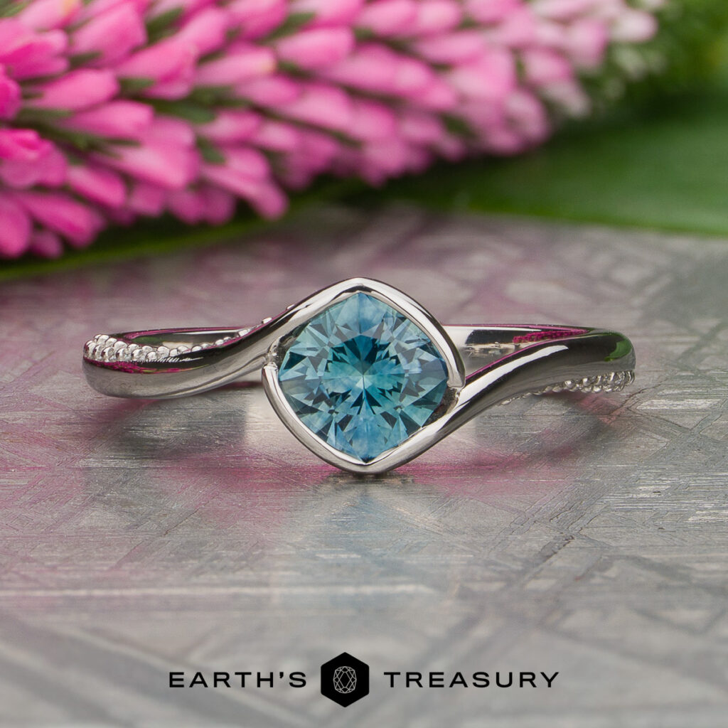 Discover the "Miller, Meghan [custom gallery]" silver ring, featuring a round blue gemstone accented by delicate side stones, beautifully showcased on a metal surface with pink flowers in the background. The image includes the "Earth's Treasury" text at the bottom.