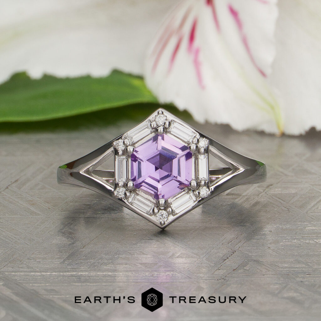 Vigh [custom gallery]: A silver ring featuring a hexagonal lavender gemstone and delicate clear accent stones, elegantly displayed on a textured surface with a flower in the background.