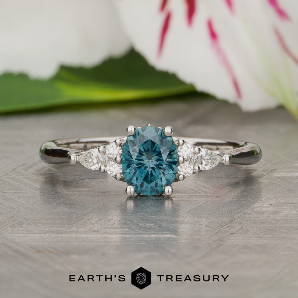 A custom silver ring from Culbert, Kristan and Ritchie, Kyle [custom gallery], featuring a round blue gemstone center flanked by two clear side stones, displayed on an ornate surface with flowers in the background and the "Earth's Treasury" logo shown below.