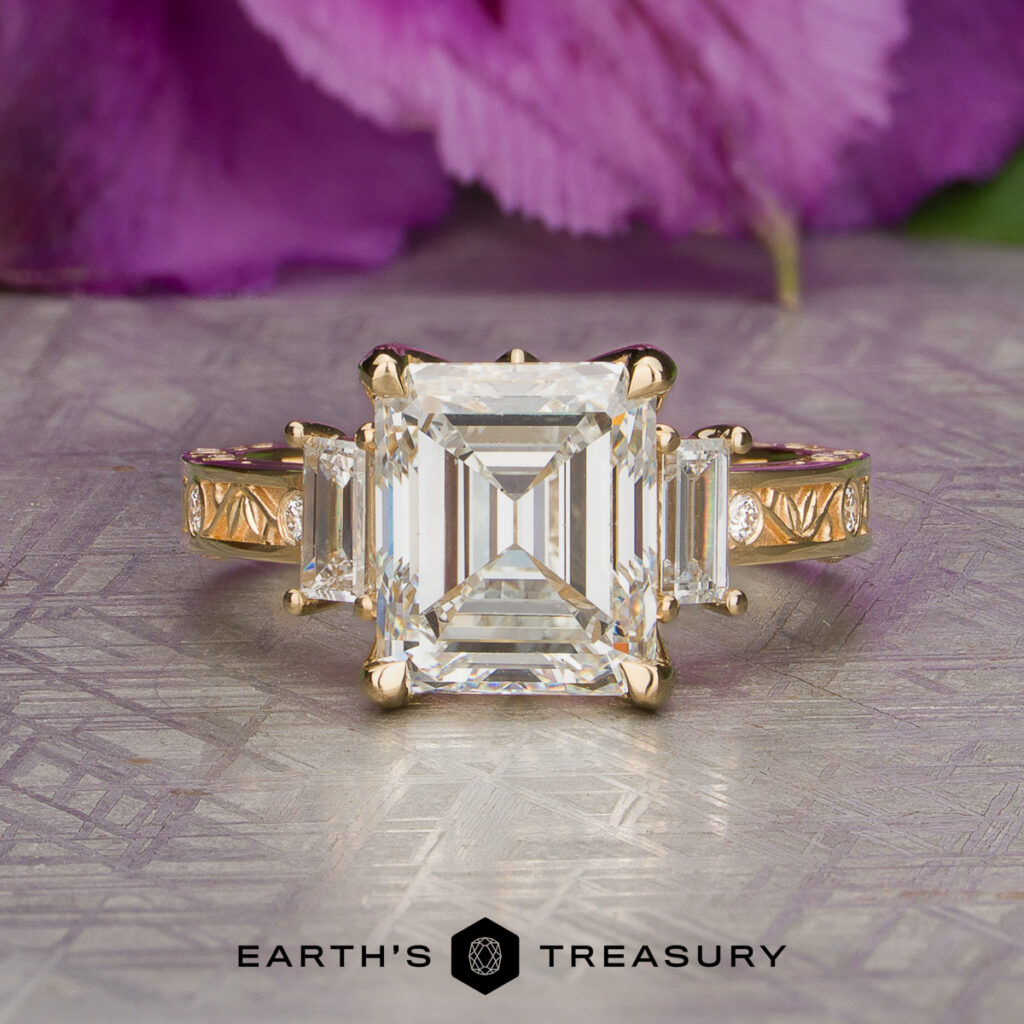 The Cohn, William [custom gallery] gold ring features a prominent rectangular gemstone at its center, accented by two smaller rectangular stones on either side, and is elegantly displayed on a textured surface with a purple flower in the background.