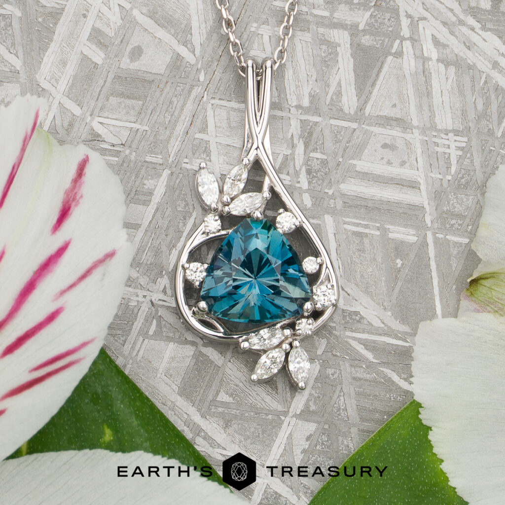 Custom pendant by Weston, Tina and Diacont, Doug featuring a silver necklace with a blue triangular gemstone accented by small clear stones, displayed on a textured surface with flower petals. The "Earth's Treasury" logo appears at the bottom.