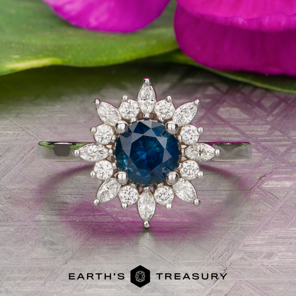 The Leung Audrey [custom gallery] silver ring features a round blue gemstone center, accented by marquise and round white stones, artfully displayed on a patterned surface with flower petals in the background.