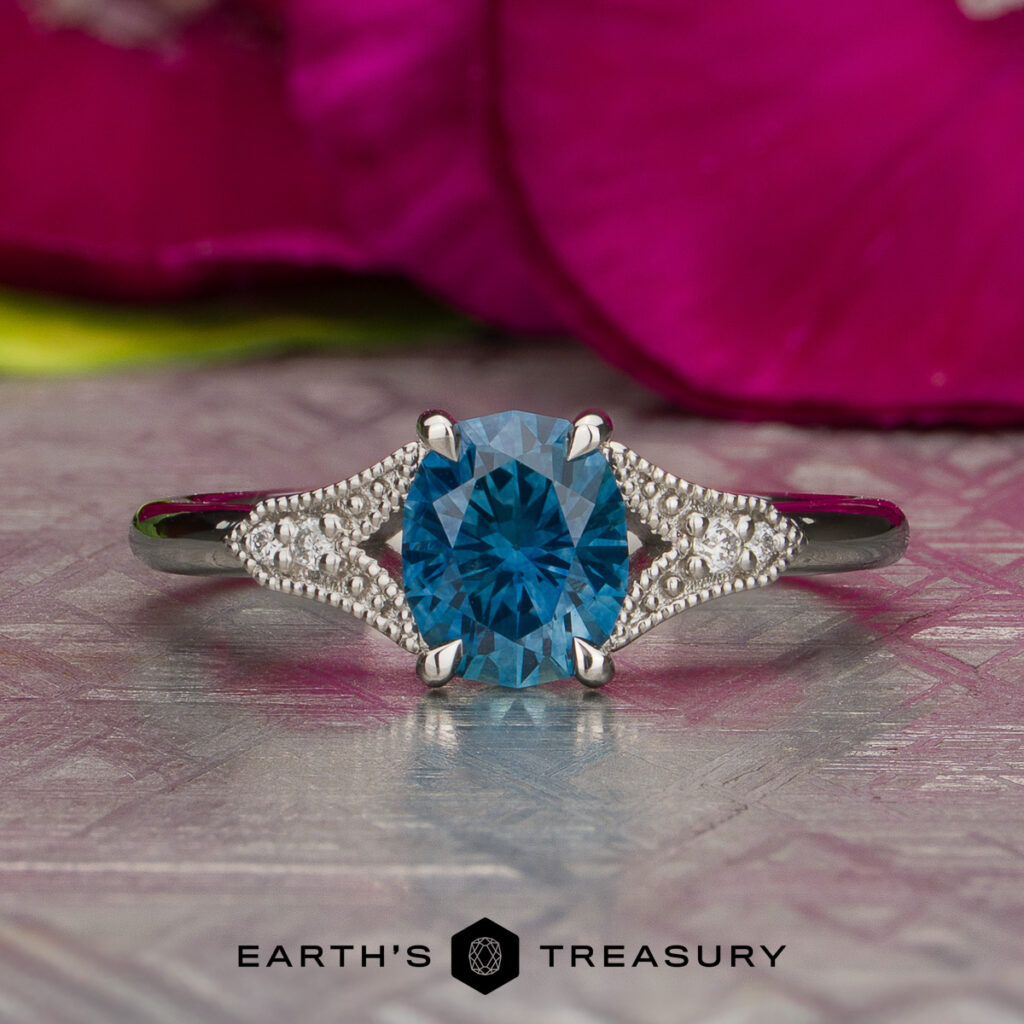 The Martin, Allena [custom gallery] ring, featuring a round blue gemstone at its center and accented by small white stones on each side, rests on a textured surface with pink petals in the background.