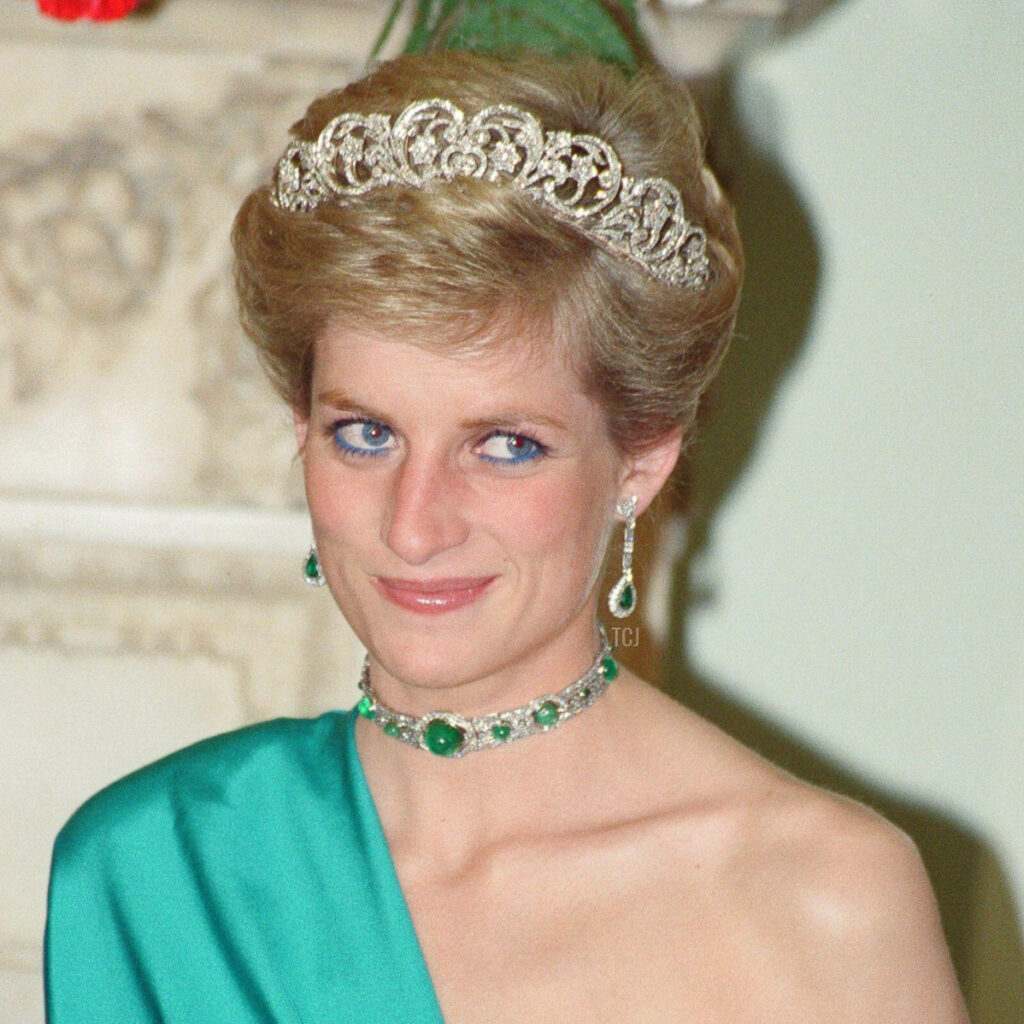 A woman wearing the diana emerald-1200-BW necklace, a jeweled tiara, and a teal dress stands indoors, looking slightly to the side and smiling softly.
