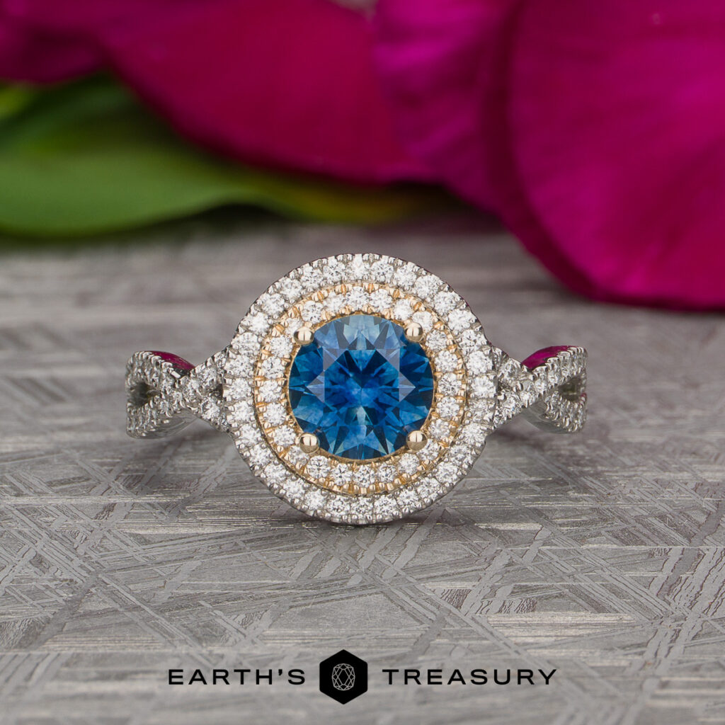 A Randall, Heather [custom gallery] ring featuring a diamond and blue gemstone with a double halo design, displayed on a textured surface with softly blurred pink flowers in the background. The Earth's Treasury logo appears at the bottom.