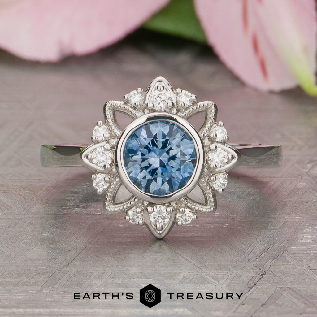 A custom gallery piece by Bowman, Luke: a silver ring featuring a central round blue gemstone encircled by sparkling white diamonds in a floral design, elegantly showcased on a textured surface.