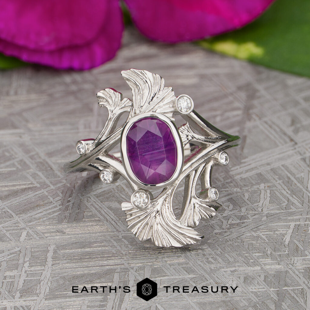 Magee, Kamille [custom gallery]: A silver ring featuring a central oval purple gemstone, accented by leaf-shaped details and small round clear stones, displayed on a textured surface with purple flowers in the background.