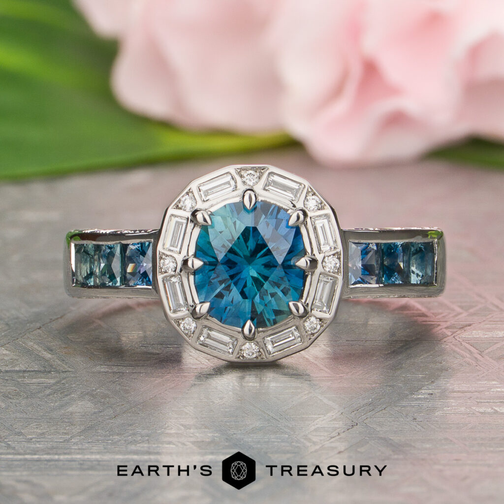 Schmidt Julie [custom gallery]: A silver ring featuring a round blue gemstone at the center, accented by both baguette and round diamonds, with square blue stones set along the band.