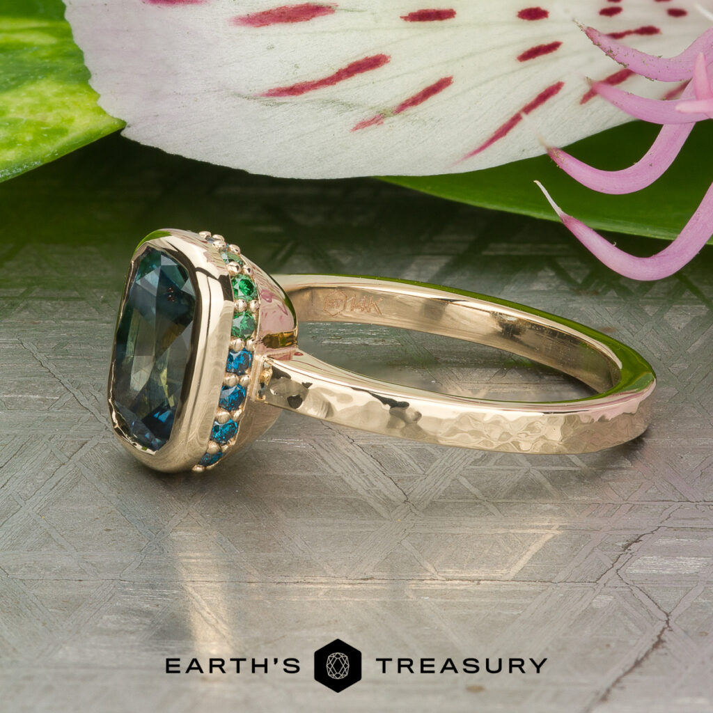 The Yu, Julie and Jorgensen, Noah [custom gallery] gold ring, featuring a striking blue gemstone accented with small green and blue stones, is displayed on a textured surface near a flower.