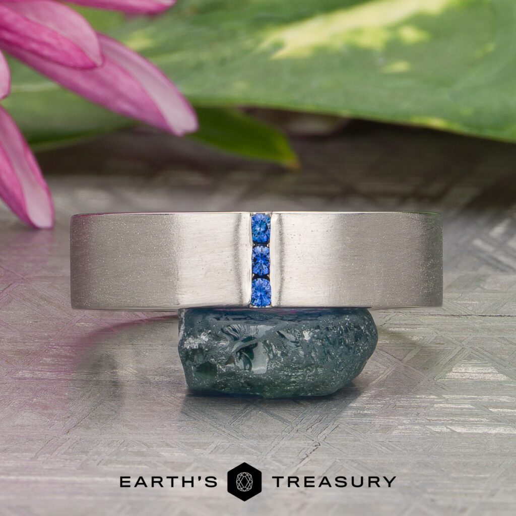 A Mathew Varkey [custom gallery] silver ring featuring three vertically-set blue stones in the center, displayed on a blue crystal with flowers and leaves in the background.