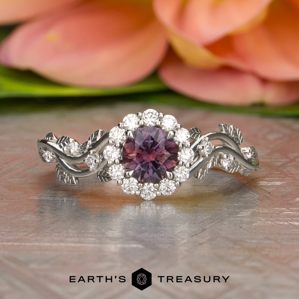 The Knutsen, Brandon [custom gallery] ring features a round purple gemstone at its center, encircled by clear stones and adorned with leaf-like accents. It is showcased on a patterned surface with blurred flowers in the background.