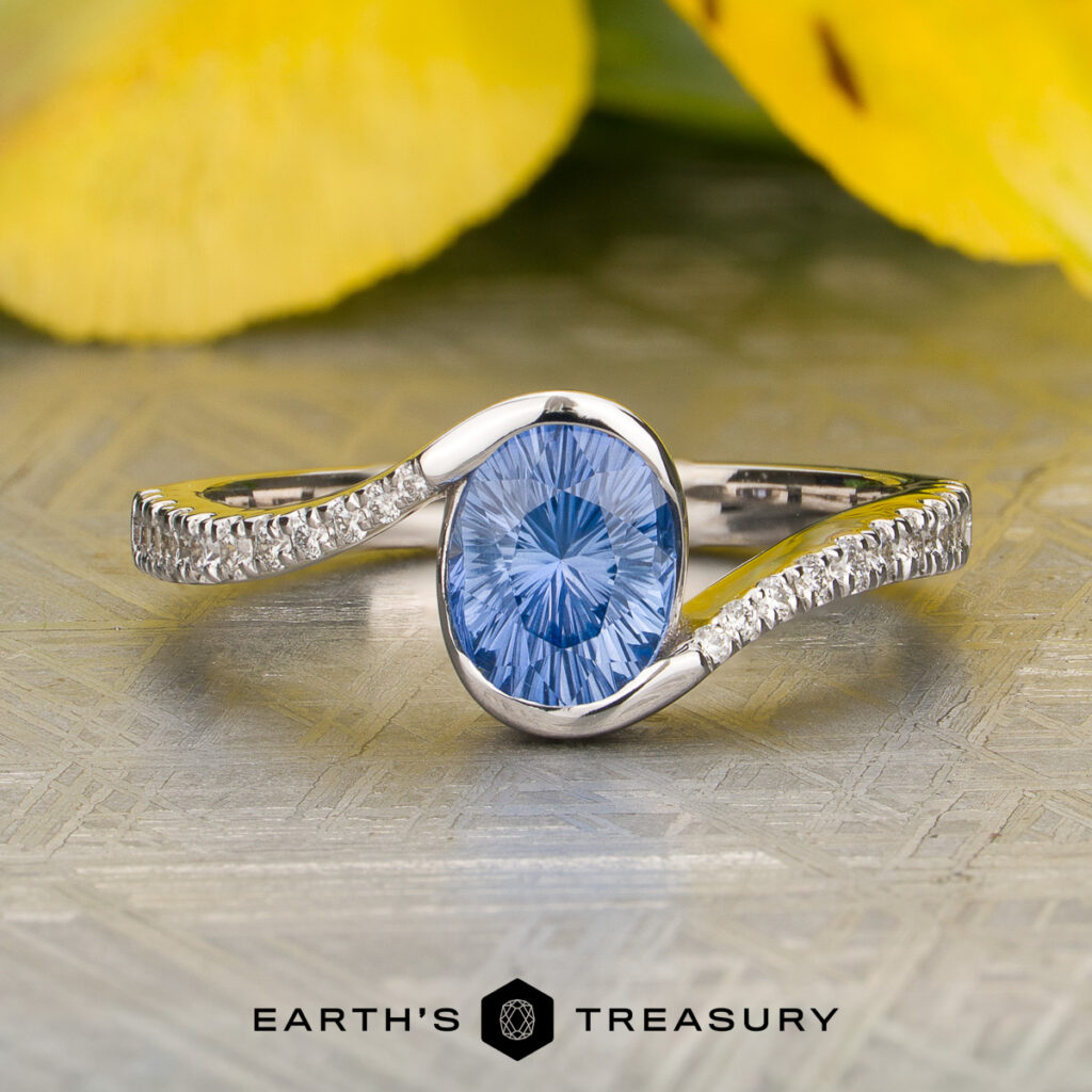 A silver "Potiker Cara" ring, featuring a blue oval gemstone flanked by small clear stones on each side, is displayed in front of yellow petals. The text at the bottom reads "Earth's Treasury.