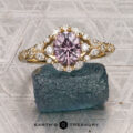 The "Eden" Halo Ring featuring a pink gemstone at its center and encircled by white diamonds is displayed atop a rough blue stone. The background shows a geometric pattern, with “Cole, Courtney and Torres, Armando 1” written below.
