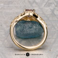 The "Eden" Halo Ring with a gemstone setting is displayed upright, resting on a blue textured stone against a patterned gray background. “EARTH’S TREASURY” is written at the bottom.