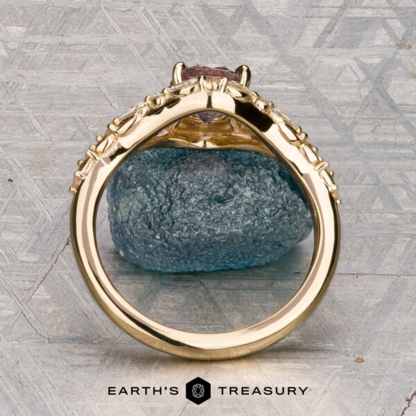 The "Eden" Halo Ring in 18K yellow gold with 0.93-carat Montana sapphire The "Eden" Halo Ring with a gemstone setting is displayed upright, resting on a blue textured stone against a patterned gray background. “EARTH’S TREASURY” is written at the bottom.