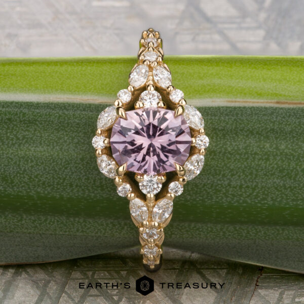 The "Eden" Halo Ring in 18K yellow gold with 0.93-carat Montana sapphire The "Eden" Halo Ring featuring a round lavender gemstone at the center, accented by marquise and round white diamonds, displayed on a green leaf.