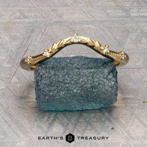The "Eden" Band featuring a gold leaf design adorned with small diamonds, is displayed upright on a rough blue stone, with the “Earth’s Treasury” logo below.