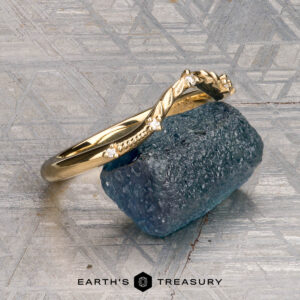 The "Eden" Band adorned with small diamonds, rests atop a rough blue gemstone. The background features a textured silver-gray surface, with the text “EARTH’S TREASURY” displayed at the bottom.