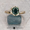 The "Cara" ring featuring an oval green gemstone and small side diamonds, displayed on a rough blue-green crystal with the “Earth’s Treasury” logo below.