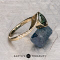 The "Cara" ring, featuring a green gemstone and small diamonds, is displayed on a rough dark blue mineral against a textured gray background. The words “EARTH’S TREASURY” are shown at the bottom.