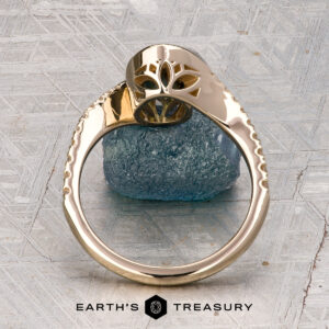 The "Cara" ring is displayed upright on a rough blue stone, featuring a decorative top with a patterned surface and the Earth’s Treasury logo beneath.