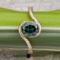 The "Cara" ring featuring a round green gemstone and a twisted band adorned with small diamonds, displayed on a green striped surface with the Earth’s Treasury logo at the bottom.