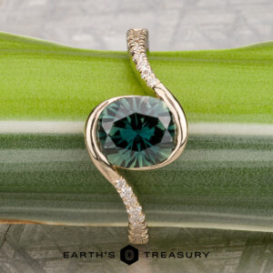 The "Cara" ring featuring a round green gemstone and a twisted band adorned with small diamonds, displayed on a green striped surface with the Earth’s Treasury logo at the bottom.