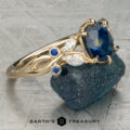 Althea: A gold ring featuring a large blue gemstone, accented by two small blue gems and two marquise-cut clear stones, displayed on a rough dark mineral with the “Earth’s Treasury” logo underneath.