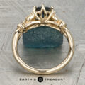Althea gold ring featuring a dark center stone secured by four prongs and flanked by two side stones, showcased upright on a blue textured rock. “Earth’s Treasury” logo appears at the bottom.