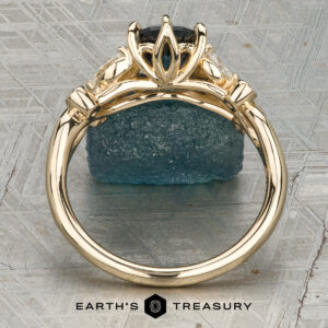 Althea gold ring featuring a dark center stone secured by four prongs and flanked by two side stones, showcased upright on a blue textured rock. “Earth’s Treasury” logo appears at the bottom.