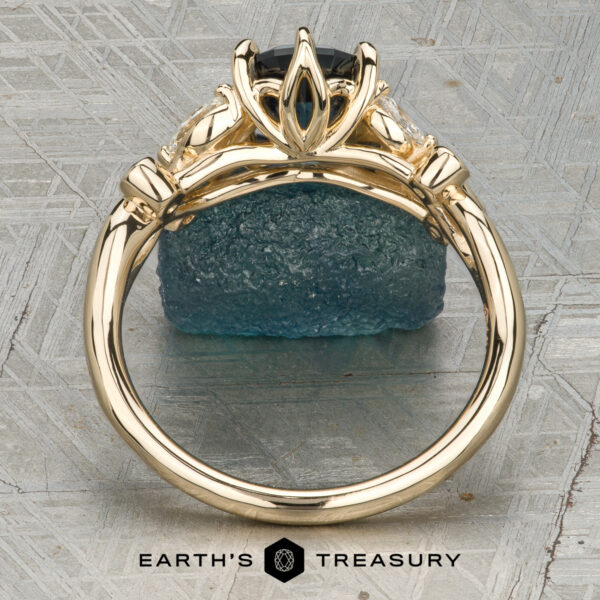 The "Althea" Ring in 14K yellow gold featuring a 1.89-Carat Royal Blue Montana Sapphire (Heated) Althea gold ring featuring a dark center stone secured by four prongs and flanked by two side stones, showcased upright on a blue textured rock. “Earth’s Treasury” logo appears at the bottom.