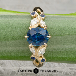 A gold “Althea” ring with an oval blue gemstone center, accented by four marquise diamonds and four small blue stones, displayed on a green stem.
