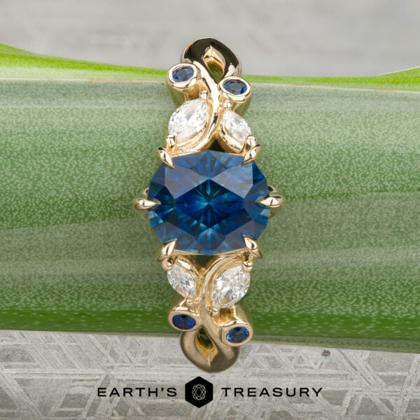 The "Althea" Ring in 14K yellow gold featuring a 1.89-Carat Royal Blue Montana Sapphire (Heated) A gold “Althea” ring with an oval blue gemstone center, accented by four marquise diamonds and four small blue stones, displayed on a green stem.