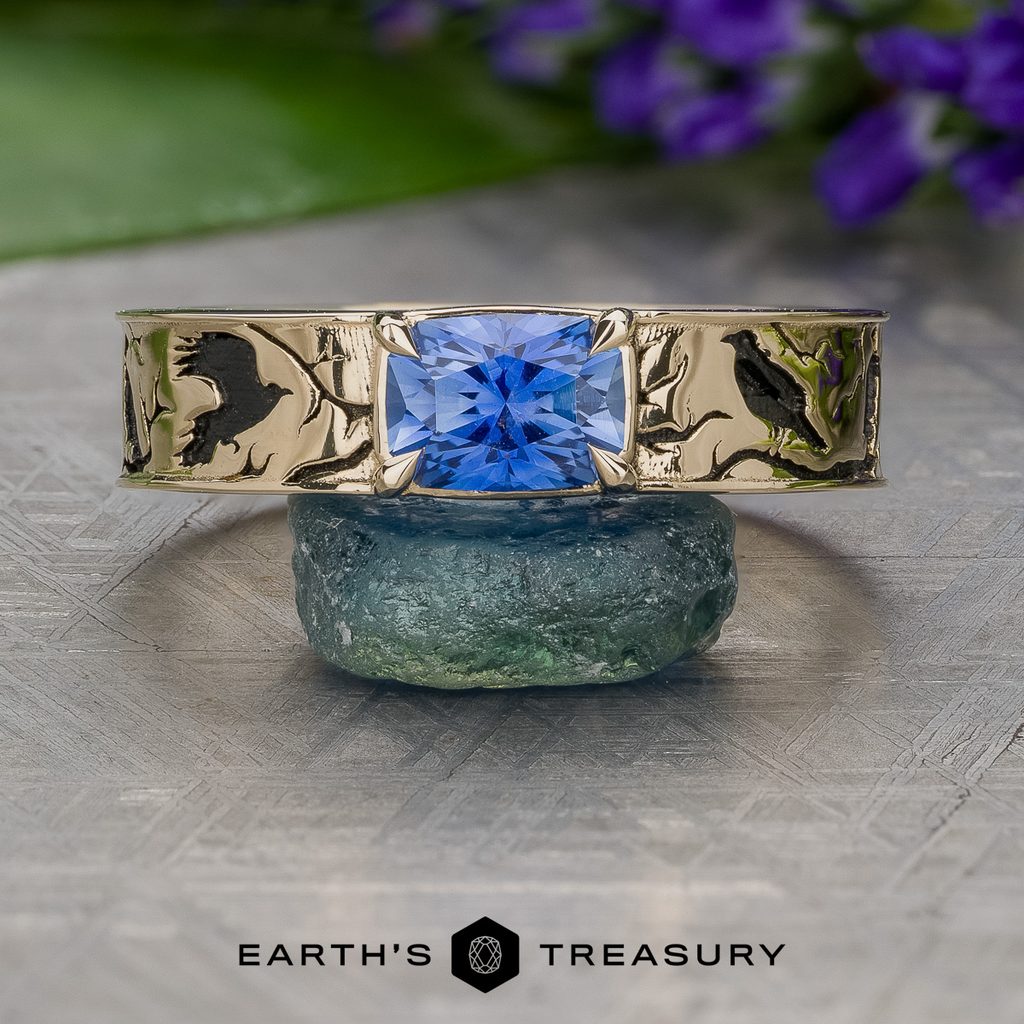 A gold ring featuring intricate raven engravings and a square blue gemstone is displayed atop a green stone, surrounded by purple flowers and leaves. Below, “Stacked from 19 images. Method=B (R=7,S=2)” is written.