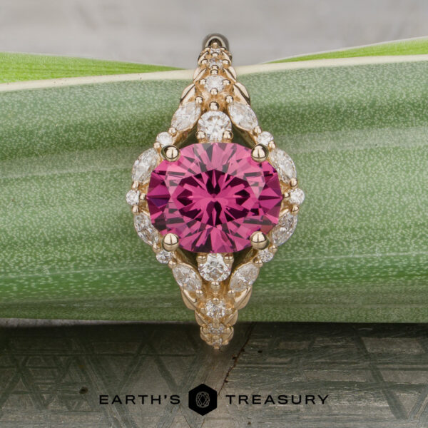 The "Eden" Halo Ring in 14K yellow gold with a 2.20ct rose pink garnet. The "Eden" Halo Ring, featuring a round pink gemstone at the center surrounded by small clear stones, displayed on a green patterned surface with “Earth’s Treasury” at the bottom.