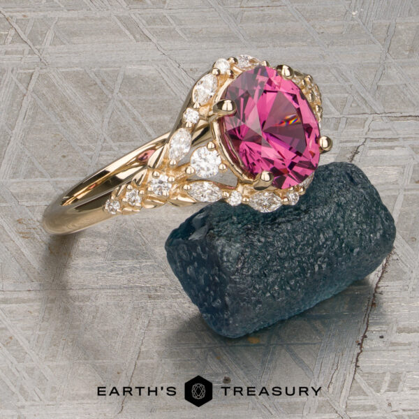 The "Eden" Halo Ring in 14K yellow gold with a 2.20ct rose pink garnet. The "Eden" Halo Ring, featuring a large pink gemstone accented with small clear stones, is displayed on a dark blue mineral sample against a textured background.