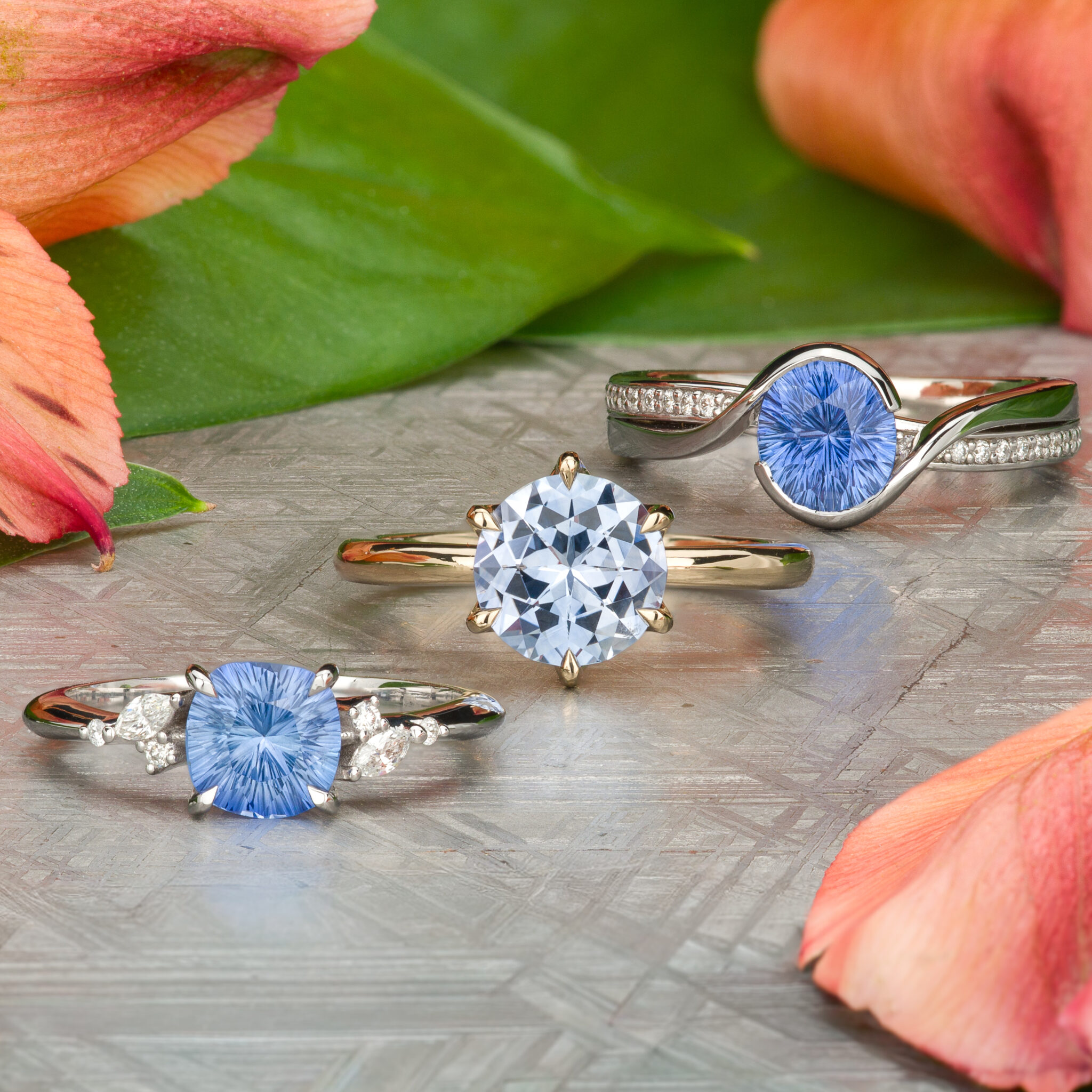 Three silver rings with light blue gemstone centers, two encircled by small diamonds, from the collection “Stacked from 71 images. Method=B (R=14,S=2)”, are displayed on a textured surface adorned with green leaves and orange flower petals.