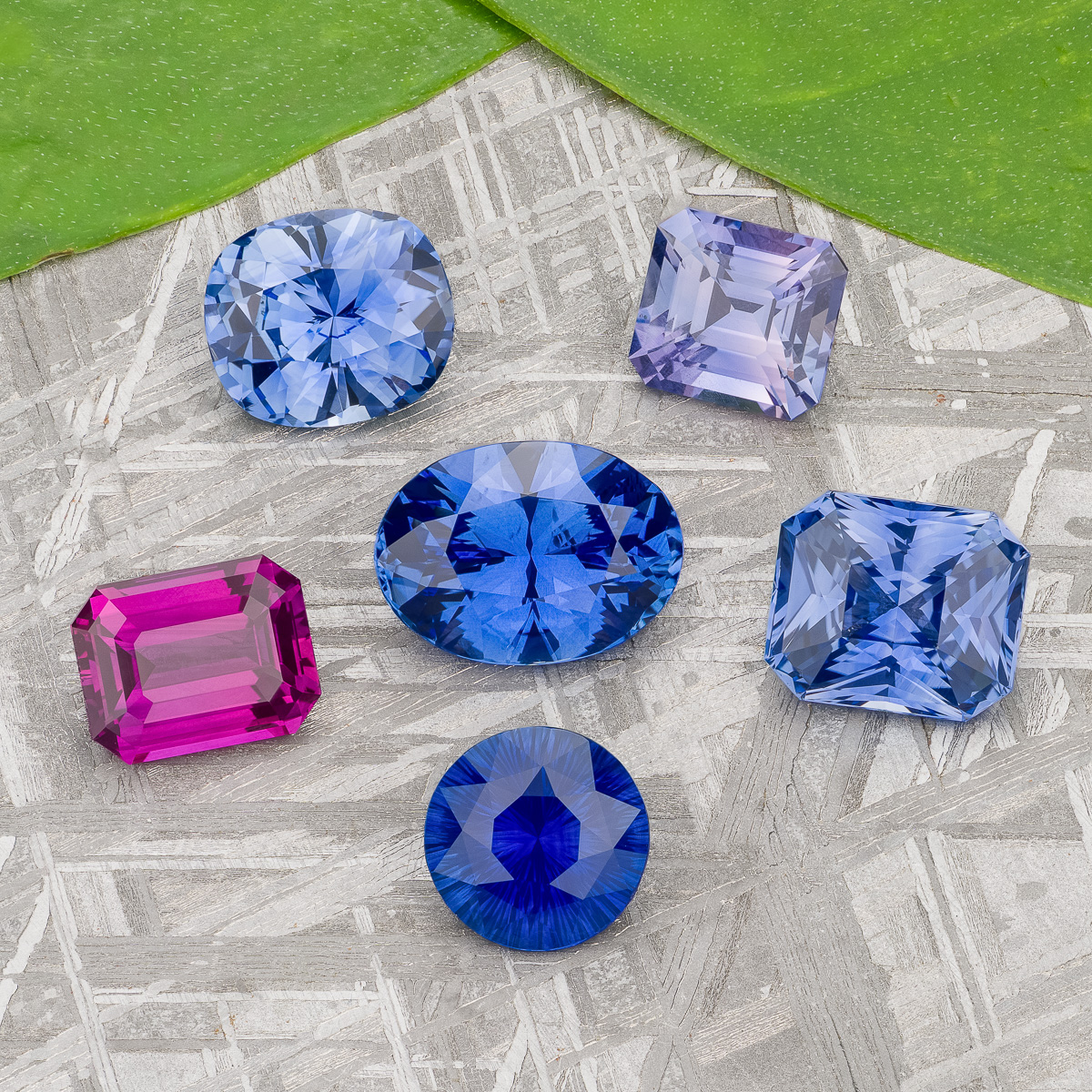 Six faceted gemstones in blue, purple, and pink hues from the “Stacked from 49 images. Method=B (R=7,S=2)” collection are arranged on a textured gray surface with two green leaves in the corner.