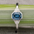 The "Everly" Three-Stone Ring in Platinum 950 with a 1.83-Carat Peacock Blue Montana Sapphire (Heated), a large square-cut blue gemstone flanked by two triangular clear stones on each side, displayed on a green and gray striped surface.
