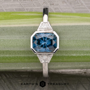 The "Everly" Three-Stone Ring in Platinum 950 with a 1.83-Carat Peacock Blue Montana Sapphire (Heated), a large square-cut blue gemstone flanked by two triangular clear stones on each side, displayed on a green and gray striped surface.