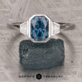 The "Everly" Three-Stone Ring in Platinum 950 with a 1.83-Carat Peacock Blue Montana Sapphire (Heated) and two white side stones, displayed on a rough blue stone with the Earth’s Treasury logo below.